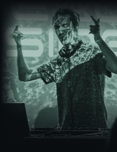 Peace Sine at the decks, arms raised, light from the screen behind him across his face